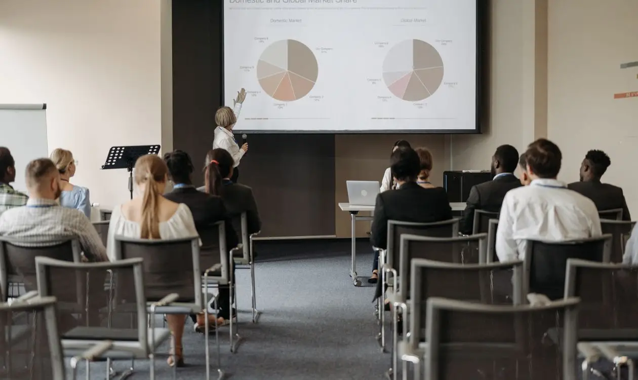 Business conference audience seated listening to a speaker presenting market data on screen
